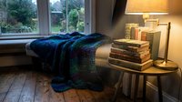 Cozy reading nook by window with stacked books, warm lamp, and blue knit blanket on wooden floor, perfect for interior design and lifestyle content.