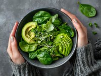 Fresh green salad bowl with sliced avocado and spinach leaves held by hands in protective gesture on dark stone background for healthy eating concept.
