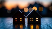 Silhouettes of miniature houses with glowing windows against blurred bokeh lights at dusk create cozy atmospheric scene. Warm illumination reflects on textured surface.