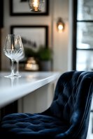 Empty crystal wine glass on white counter with luxurious navy blue velvet tufted chair and warm ambient lighting creating elegant restaurant atmosphere.