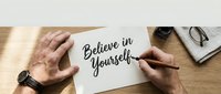 Male hands writing motivational quote Believe in Yourself on white paper at wooden desk with notebook and glasses for inspiration and self-development.