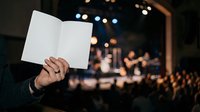 Person holding blank white open brochure or pamphlet at corporate event with blurred audience and stage lights in background for marketing and business communications.