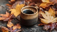 Rustic ceramic cup of hot coffee surrounded by colorful autumn maple leaves on wooden surface, creating cozy fall atmosphere for seasonal beverage enjoyment.