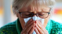 Senior woman in glasses blowing nose into tissue, showing cold or flu symptoms. Wearing green sweater against blurred background, demonstrating proper hygiene during illness.