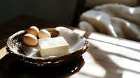 Fresh farm eggs and block of tofu on rustic ceramic plate in morning sunlight with dramatic shadows, minimalist food photography composition.