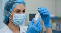 Female healthcare worker in surgical mask and cap holding medical vial in laboratory setting for pharmaceutical research and vaccine development.