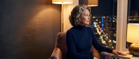 Mature Caucasian businesswoman in navy turtleneck sitting thoughtfully by window with city lights at night in modern office or hotel room interior.