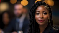 Young African American woman with long braided hair wearing professional black attire, looking at camera with confident expression in blurred office environment with colleagues.