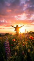 Woman with arms raised celebrating freedom in wildflower meadow at golden sunset with dramatic purple and orange sky for wellness and inspiration content.