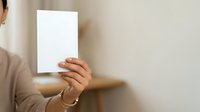 Young woman with medium skin tone holding blank white card or paper against neutral beige wall, suitable for mockups, advertising campaigns, and promotional materials.