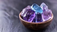 Raw amethyst and blue topaz crystals in rustic wooden bowl against dark background, healing stones and minerals with mystical purple and blue glow.