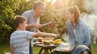 Middle-aged Caucasian man and young Caucasian woman with a boy enjoying outdoor meal together in sunny garden setting with food on table and green foliage background.