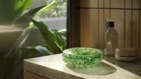 Green glass ashtray on marble counter near window with soft natural lighting and plant leaves, creating serene minimalist composition with glass bottle in background.