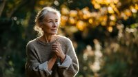 Senior woman with gray hair in cozy sweater enjoying peaceful moment in autumn forest with golden bokeh background at sunset.