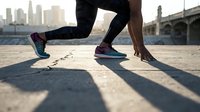 Athletic runner in starting position on urban pavement, wearing colorful sneakers and patterned leggings, city skyline in background at sunrise.