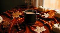 Steaming mug of hot coffee on rustic orange blanket with autumn leaves, cinnamon sticks, and white pumpkins near window.