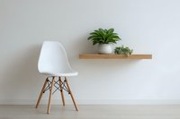 Modern white chair with wooden legs next to floating shelf displaying green plant and decorative items against minimalist wall interior design.