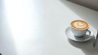Cup of cappuccino with latte art heart pattern on white table with natural sunlight creating soft shadows for cafe and coffee shop concepts.
