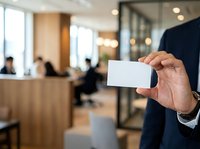 Businessman in dark suit holding blank white business card in modern office lobby with blurred colleagues and reception desk in background for corporate branding and networking.