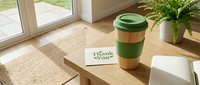 Eco-friendly reusable bamboo coffee cup with green lid beside a thank you card on a wooden table in a bright, cozy home interior.