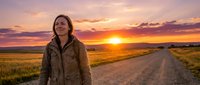 Woman in brown jacket standing on rural dirt road at golden hour sunset with expansive grassland and dramatic sky, suitable for travel, lifestyle, and outdoor adventure content.