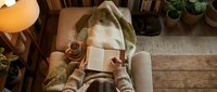 Young woman relaxing on cozy sofa reading book with warm tea, comfortable home interior with plants and wooden floors for lifestyle content.
