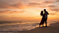 Romantic couple silhouette dancing on beach at sunset with golden sky and ocean waves creating intimate moment for wedding anniversary celebration.
