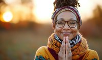 African American woman wearing glasses, colorful headwrap and winter scarf smiling with hands pressed together in prayer position against warm sunset background.