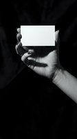 Female hand with dark nail polish holding blank white card on black fabric background, monochrome mockup for branding and design.