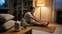 Young woman practicing yoga stretches on mat in cozy living room with warm lamp lighting and essential oil diffuser for home wellness routine.