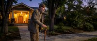 Senior Caucasian man with walking cane standing in residential driveway during twilight hours with illuminated house and mature trees in background.