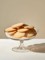 Fresh butter cookies arranged on elegant glass cake stand against neutral beige background with soft shadows and minimalist composition.