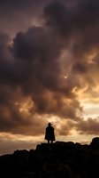 Silhouette of lone person standing on rocky cliff against dramatic stormy sky with golden sunset clouds for adventure and solitude concepts.