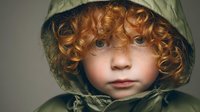 Portrait of young child with curly ginger hair and blue eyes wearing green hooded raincoat, looking directly at camera with innocent expression.