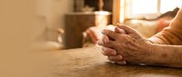 Elderly hands clasped in prayer on wooden table with warm sunlight streaming through window creating peaceful spiritual moment for meditation.