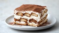 Classic tiramisu dessert with layers of coffee-soaked ladyfingers and mascarpone cream, dusted with cocoa powder on white plate.