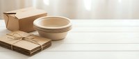 Eco-friendly packaging materials including cardboard boxes, kraft paper bundles and biodegradable bowls on white wooden surface for sustainable shipping.