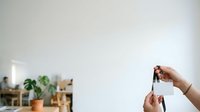 Person holding house keys in hand against blurred home interior background with plants and shelving, representing real estate, property ownership, moving, and home purchase concepts.