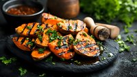 Grilled sweet potato slices garnished with fresh herbs and sesame seeds on black stone plate, moody food photography with natural lighting and rustic styling.
