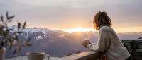 Young woman with long brown hair, wearing a beige knit sweater, holding a warm beverage while gazing at snow-capped mountains during golden hour sunrise or sunset. Peaceful winter morning scene for lifestyle and wellness content.
