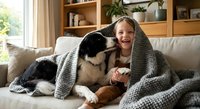 Young girl cuddling with border collie dog under cozy knit blanket on living room sofa. Warm family bonding moment at home with pet companion and natural lighting.