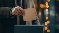 Person casting vote in ballot box during election with bokeh lights in background, creating atmospheric democratic moment. Cropped view focuses on hand and ballot.