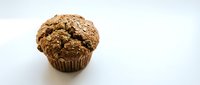 Freshly baked chocolate chip muffin with crumbly topping in brown paper wrapper on white background, suitable for bakery, food, and culinary content.