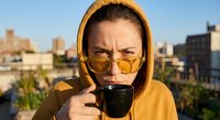 Young Caucasian woman in yellow hoodie and sunglasses drinking coffee on urban rooftop terrace during golden hour with city skyline background.