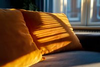 Sunlight through window blinds casting dramatic shadows on orange pillows on dark sofa, creating cozy morning atmosphere with warm light patterns.