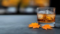 Glass of whiskey with ice cubes on dark surface next to autumn maple leaves with blurred warm kitchen background for seasonal cocktail concepts.