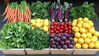 Fresh organic produce display with vibrant carrots, leafy greens, purple beets, tomatoes, and citrus fruits arranged in wooden crates at farmers market or grocery store.