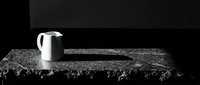 White ceramic pitcher on textured gray fabric with fringed edges against dark background, suitable for still life and product photography.