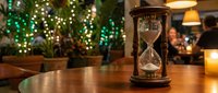 Vintage hourglass timer on wooden table in cozy restaurant with warm lighting and festive green lights creating atmospheric dining ambiance.