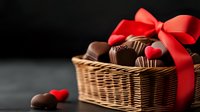 Dark chocolate pralines and truffles in wicker basket decorated with red satin bow and heart shapes on black background, romantic gift for Valentine's Day celebration.